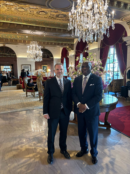 2 men in black suits standing under a chandelier in a large reception area