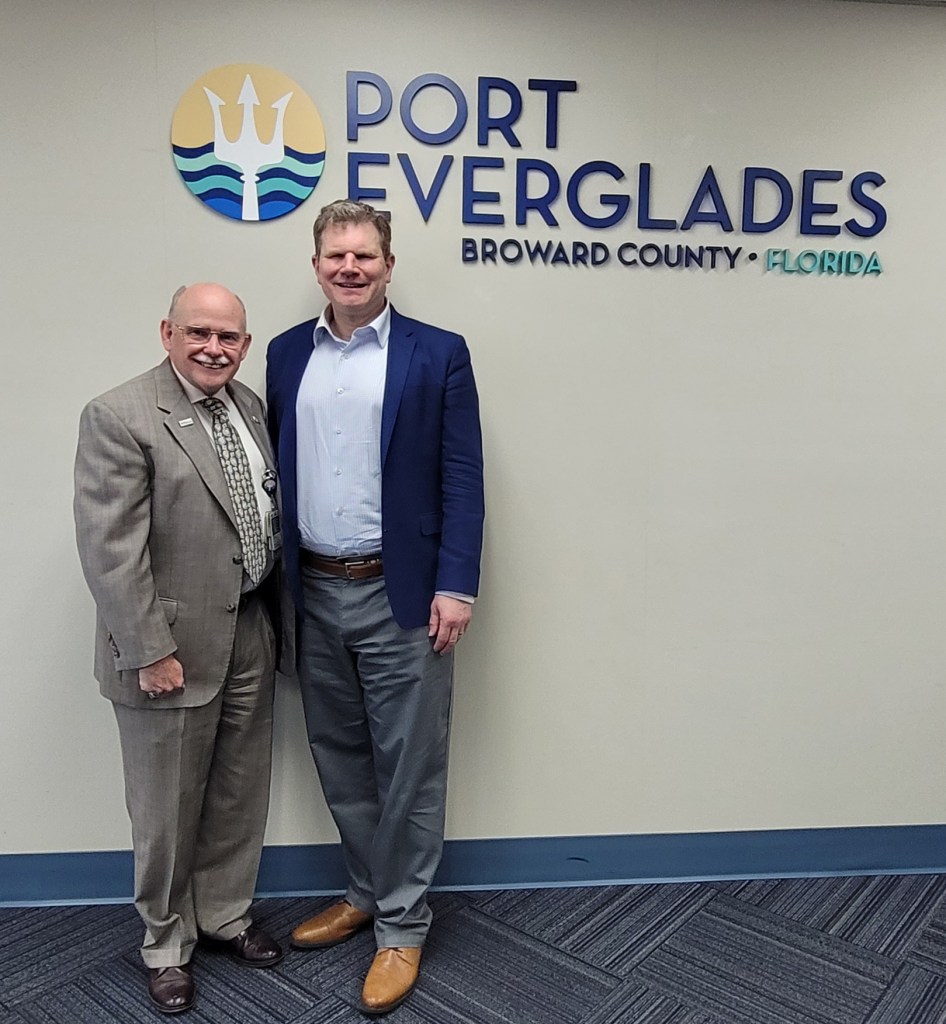 two men in suits standing against a wall under the sign and logo of the Port of Everglades; Broward County Florida
