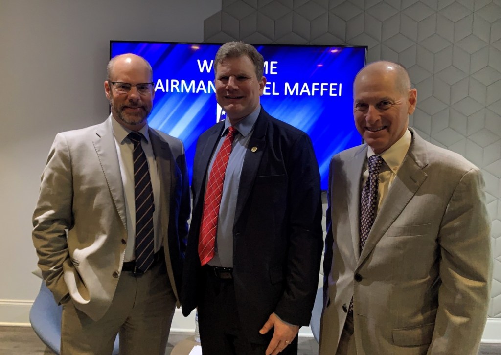 three men in suits standing facing the camera in front of a large tv screen with the name of the center man on it