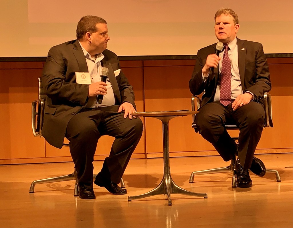 Two men in suits seated in chairs on a stage. Both men have microphones. One man is talking while the other listens.